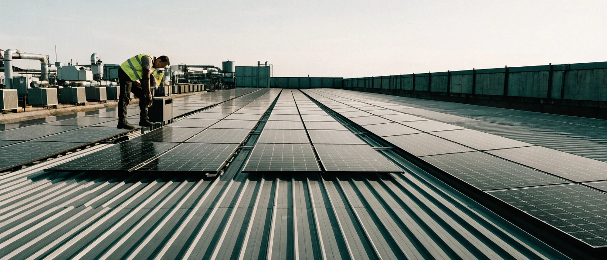 An electrician in a high-visibility vest working at the edge of a large rooftop solar array on a factory roof, roof plant behind.