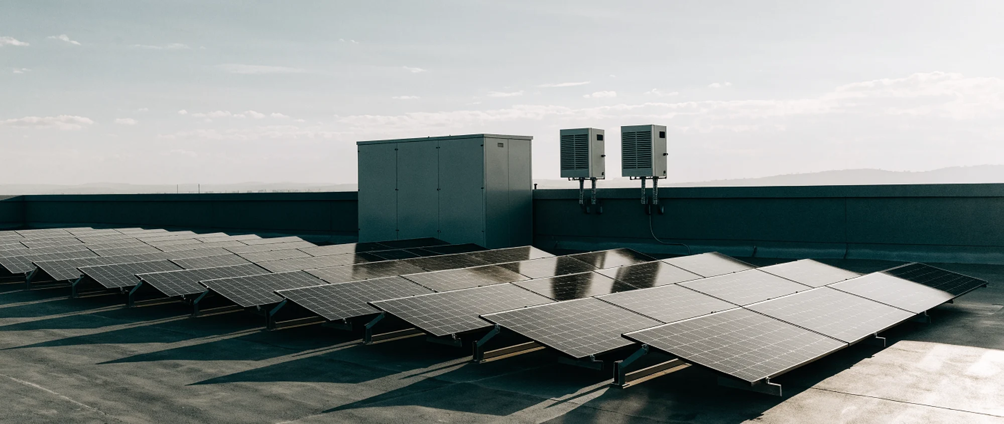 A rooftop solar array in the foreground with a battery cabinet and two inverters standing at the parapet edge behind it.