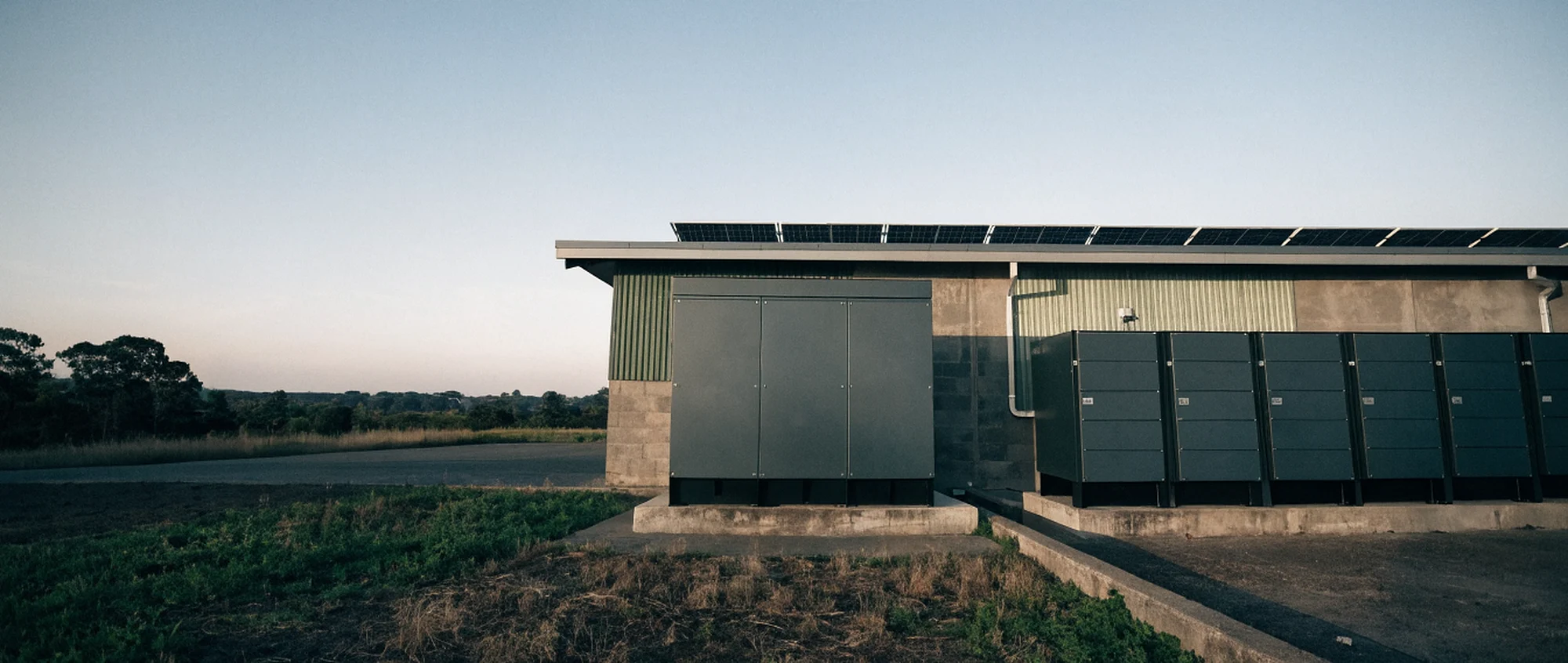 A battery energy storage cabinet and inverter bank on a concrete pad outside a light industrial building, with a rooftop array along the roofline behind.