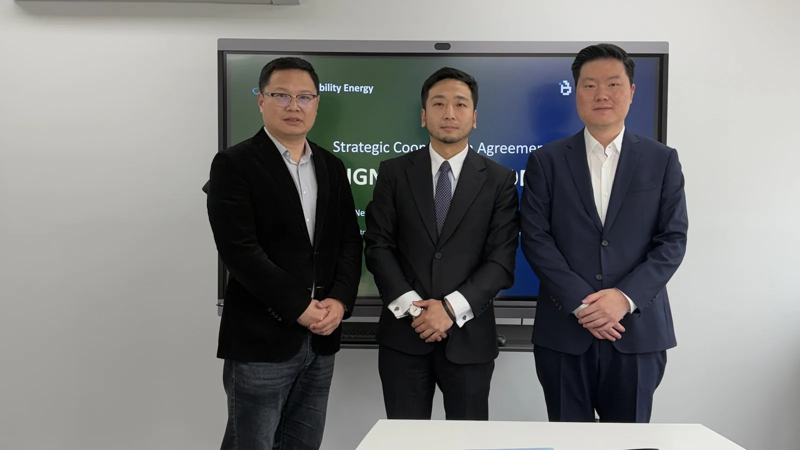 Three representatives of New Ability Energy and BitWatt Energy standing side by side in a meeting room, in front of a screen reading Strategic Cooperation Agreement Signing.