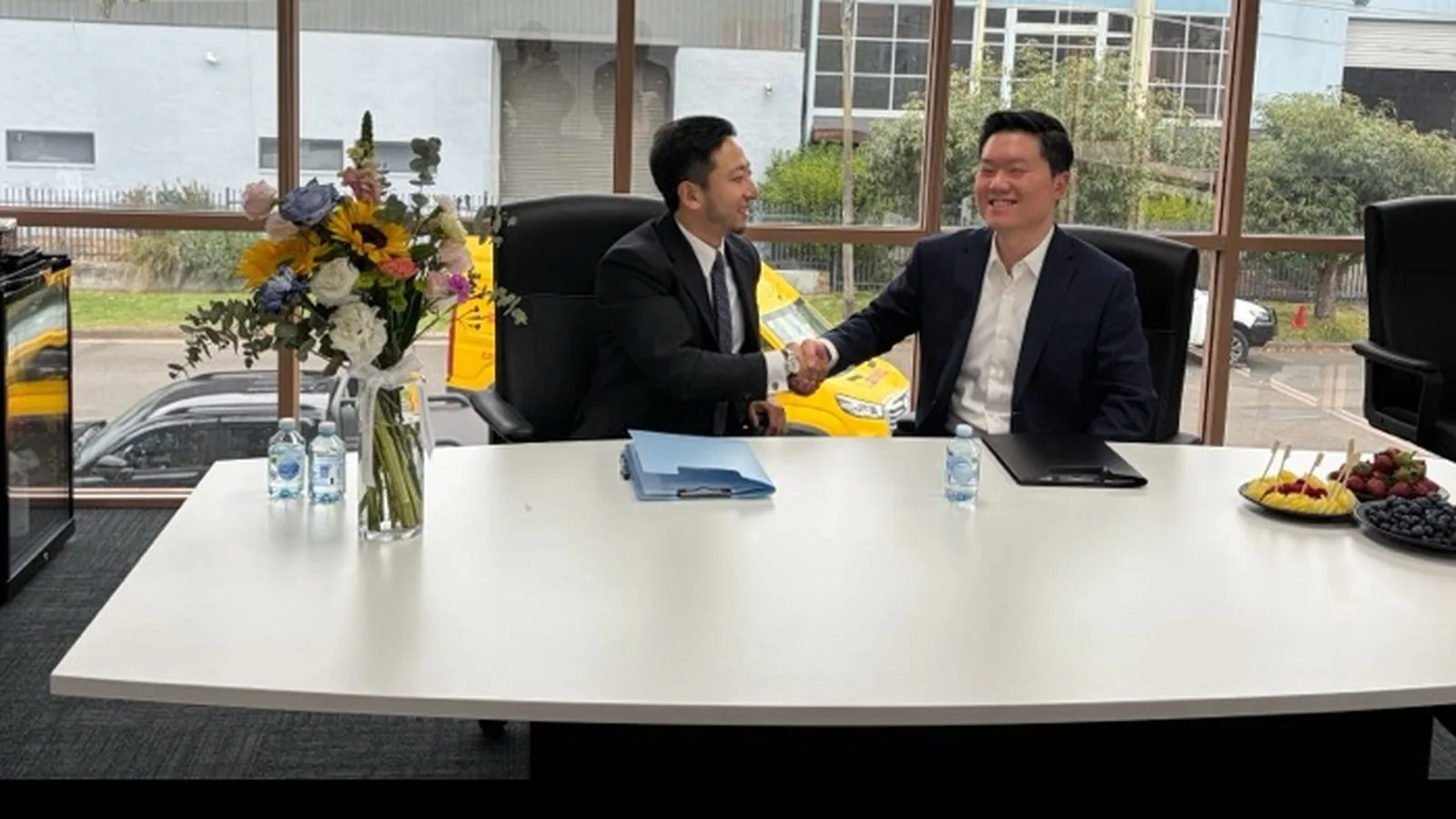 Two representatives shaking hands across a boardroom table after signing the agreement, with the signed folders in front of them.