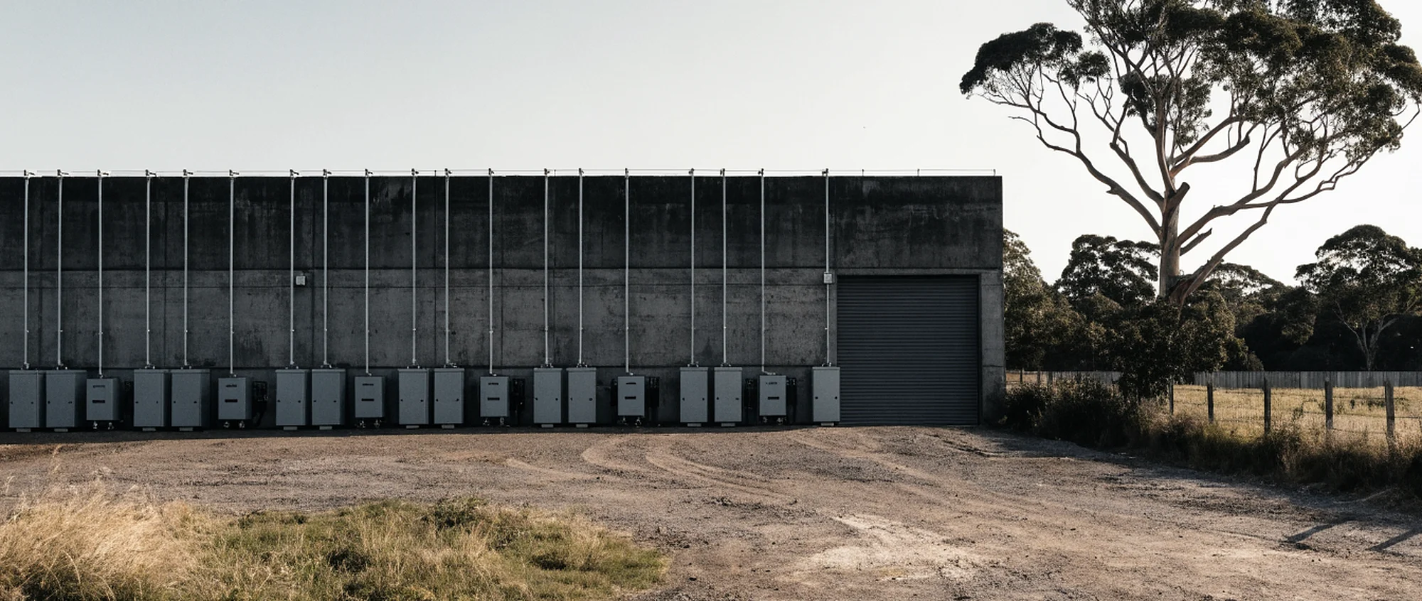 A row of battery enclosures mounted along the wall of a tilt-slab industrial unit, in a gravel service yard with a stringybark at the fence line.
