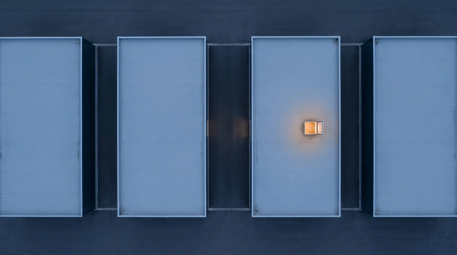 A view straight down at dusk on four identical bare commercial roofs in a row, with one small roof hatch lit on the third.