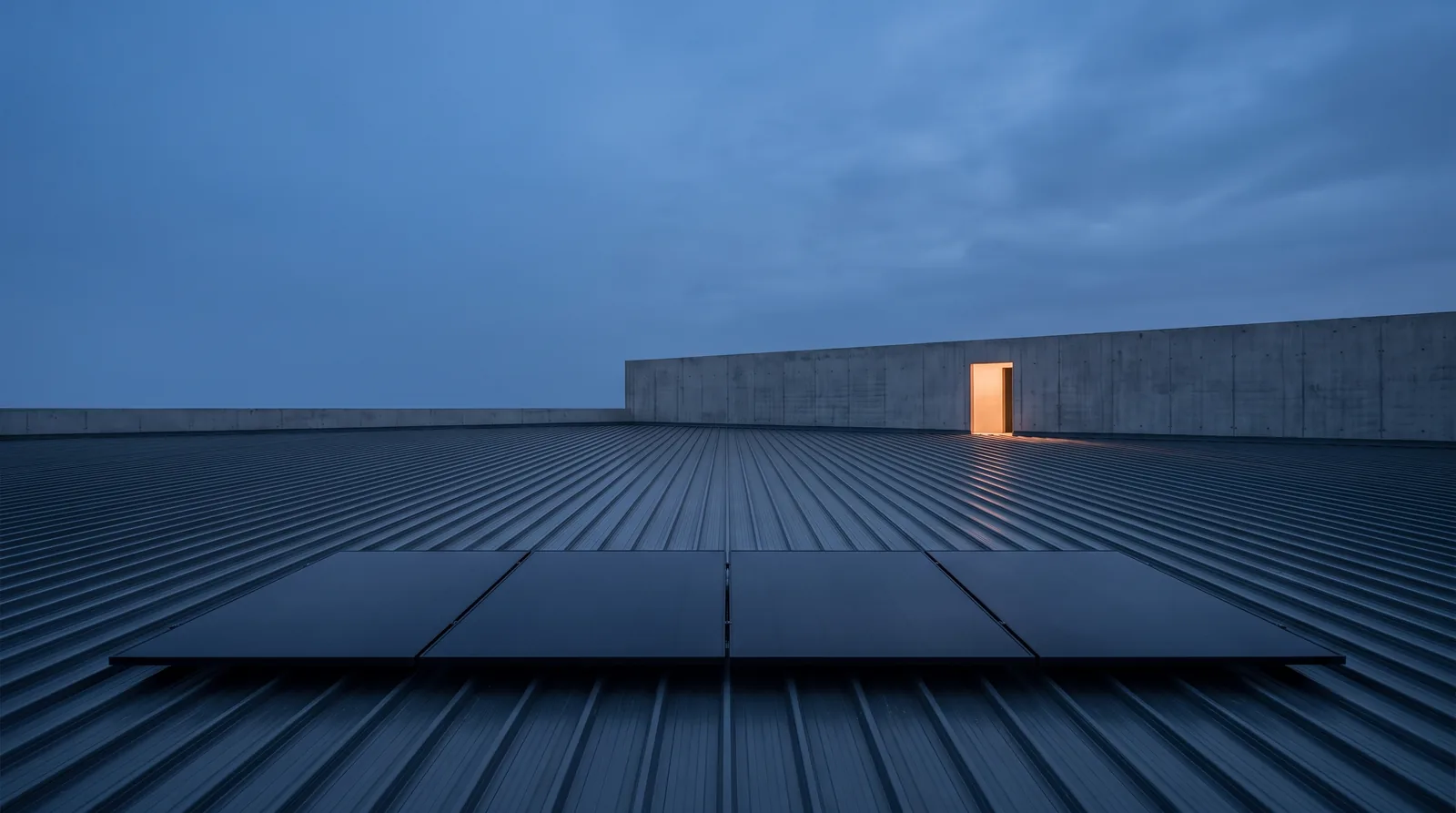 A single short row of solar panels at the near edge of an otherwise bare steel roof deck at dusk, with a lit doorway in the far parapet.
