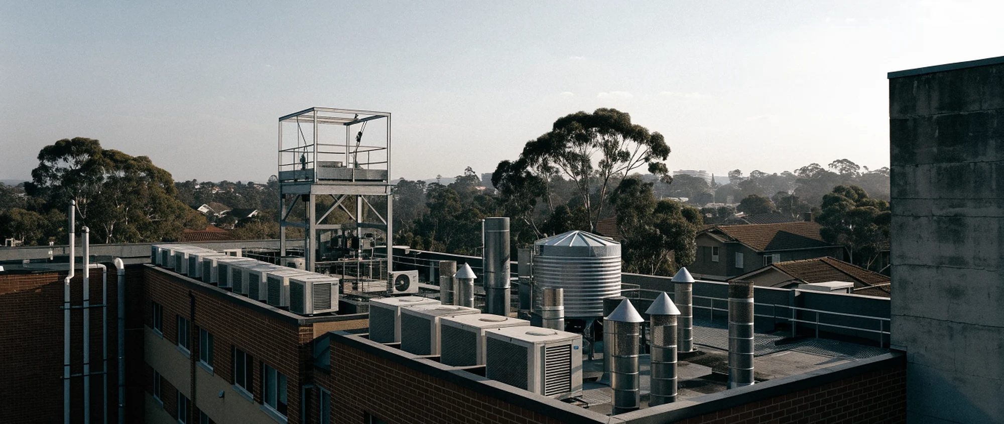 The roof of a brick apartment building, crowded with air conditioning plant, vent stacks, a water tank and a lift overrun, with suburban rooftops and gums beyond.