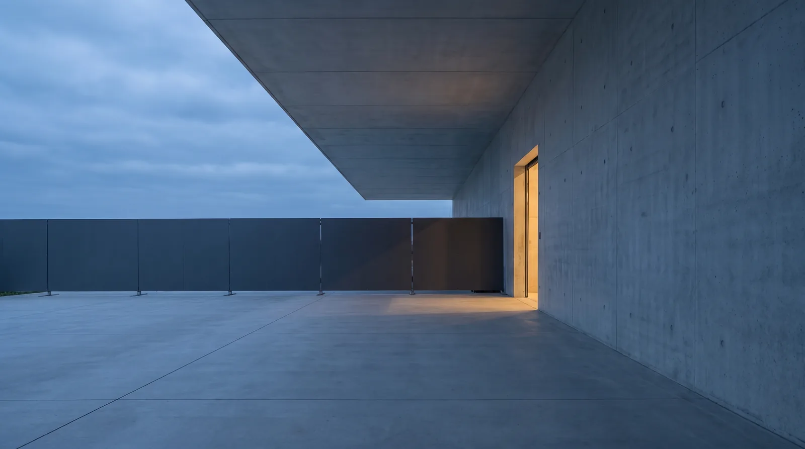 A covered concrete walkway at dusk beside a lit doorway, with a line of charcoal temporary barrier panels closing off the space beyond it.