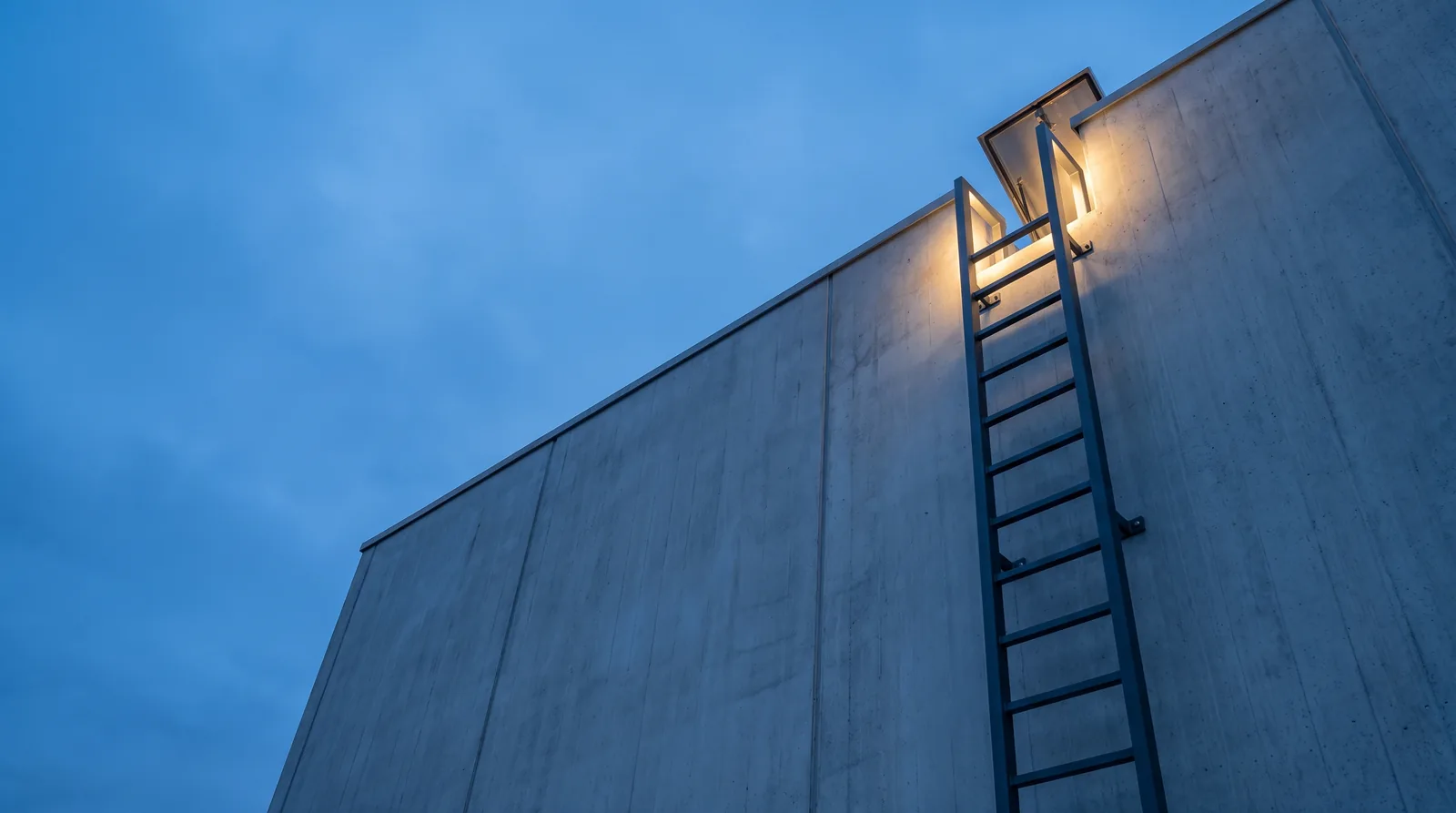 A fixed steel ladder rising up a concrete wall at dusk to an open roof hatch, with warm light spilling from the hatch.