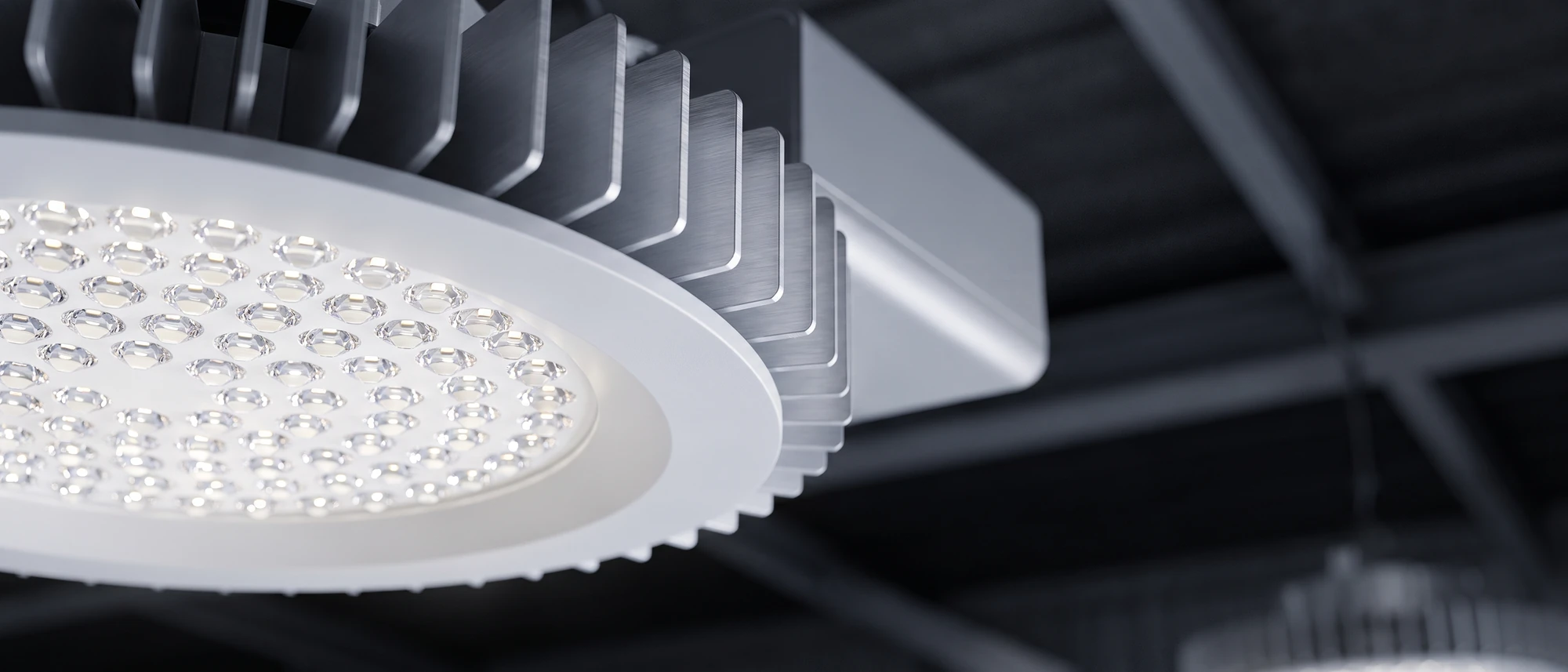 Close-up of a commercial LED high-bay luminaire in service, its lens array and aluminium heatsink fins sharp against an out-of-focus warehouse ceiling.