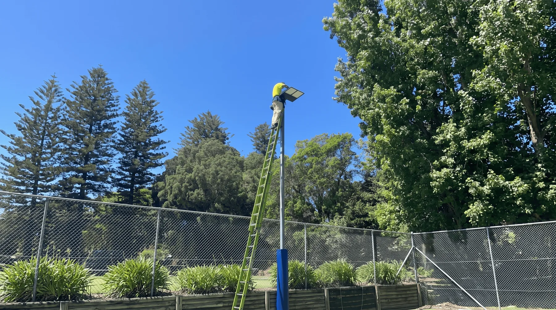 An electrician on an extension ladder fitting an LED floodlight to a pole above a fenced sports court.