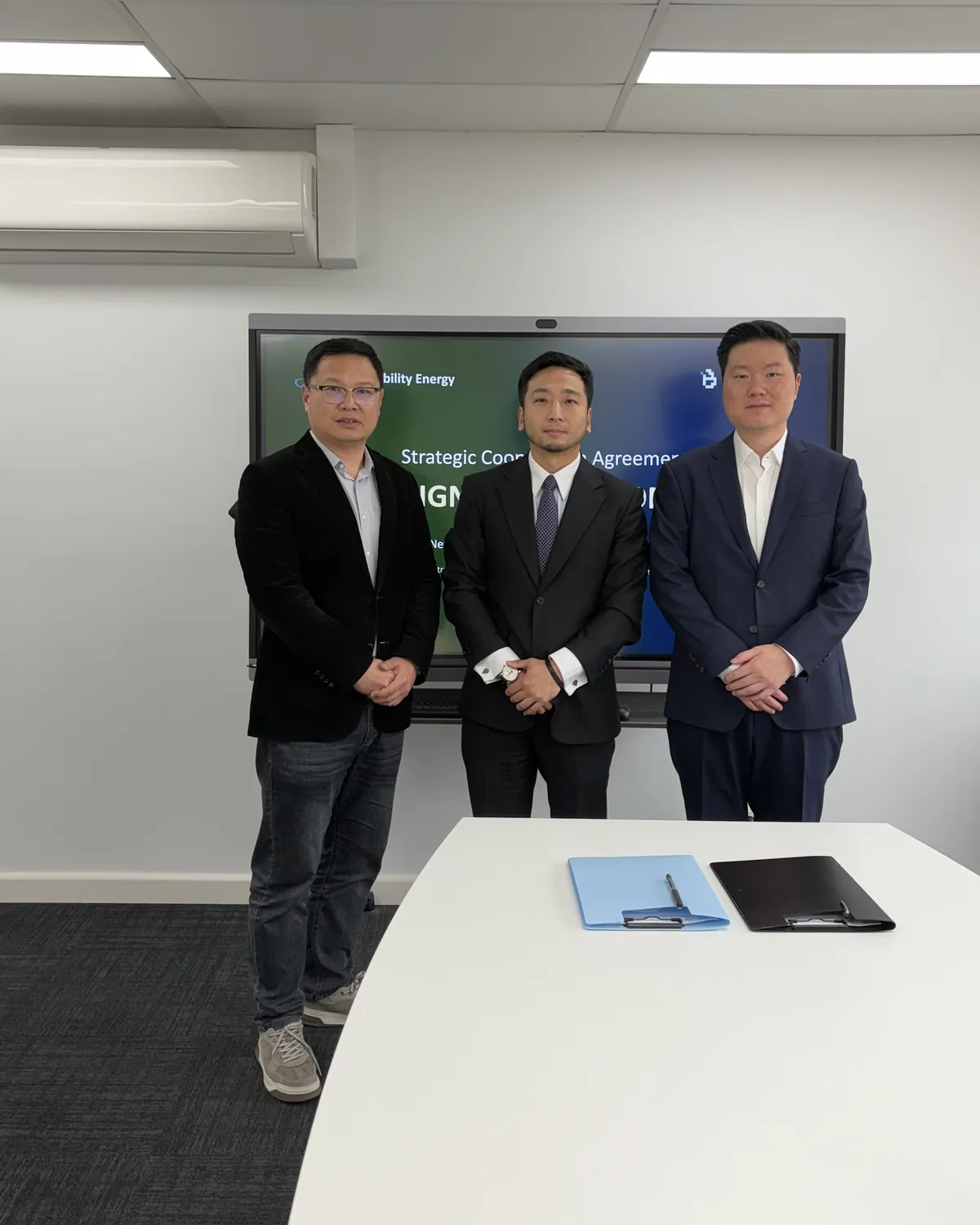 Three representatives of New Ability Energy and BitWatt Energy standing in a meeting room in front of a screen reading Strategic Cooperation Agreement, the two signed folders on the table.