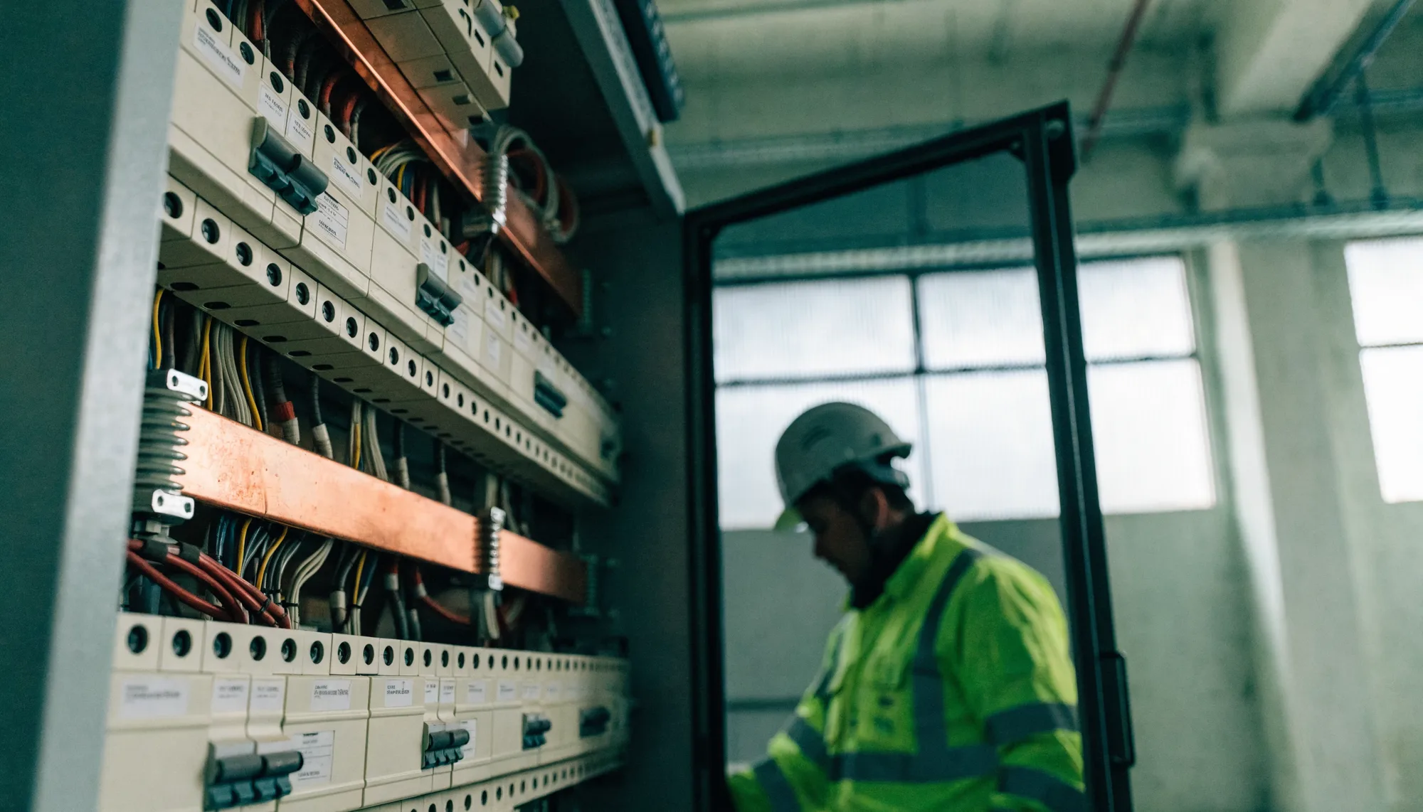 An open commercial switchboard with rows of labelled circuit breakers and copper busbar, an electrician in high-visibility workwear working at it.